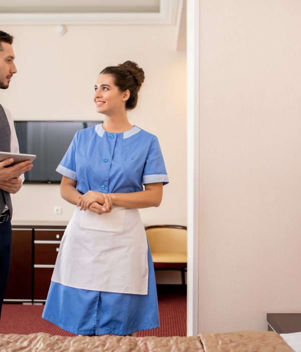 Cheerful young room maid in uniform talking to elegant porter with touchpad